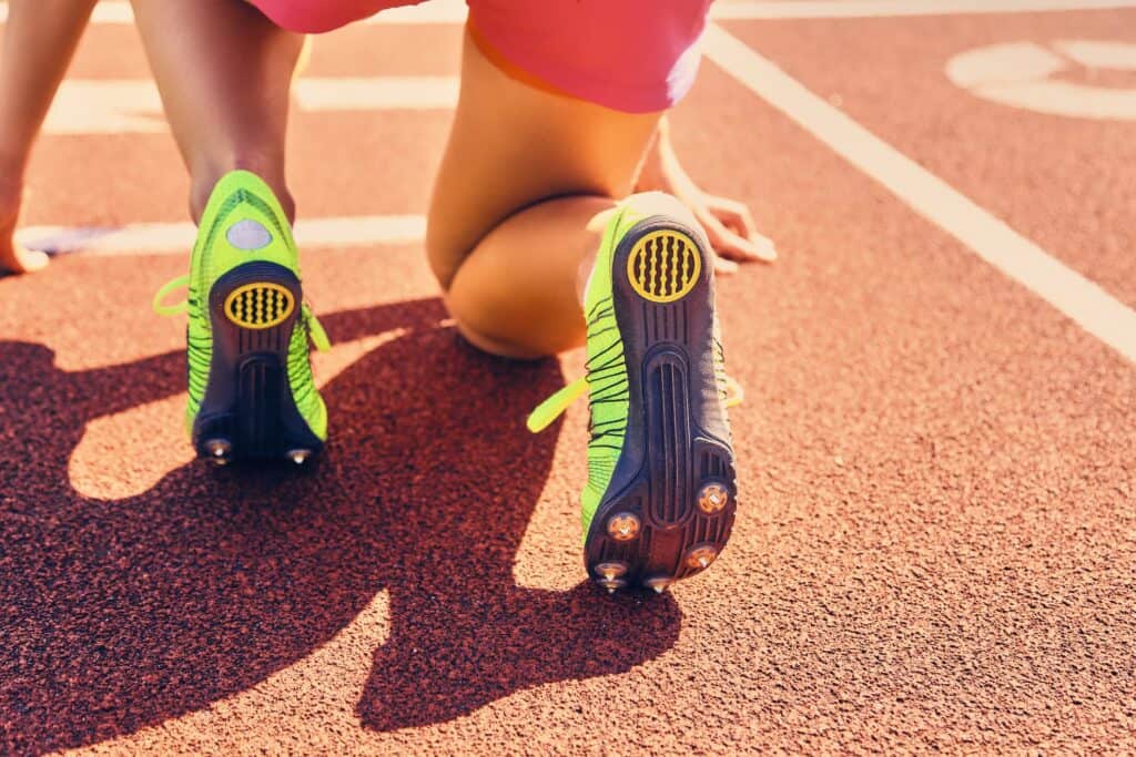 Primer plano de un corredor en la línea de salida de una pista de atletismo, con zapatillas verdes brillantes con clavos, en posición de salida agachado, listo para esprintar, atento a las lesiones y a los avances de la ortopedia deportiva para rendir al máximo.
