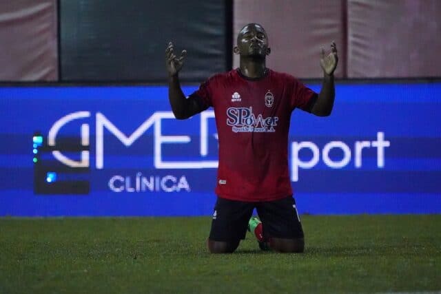 Un jugador de fútbol con camiseta roja se arrodilla en el campo con los ojos cerrados y las manos levantadas, como si estuviera celebrando o dando las gracias, tal vez aliviado tras haber superado unas lesiones. Al fondo se ve un cartel publicitario azul de Clínica GMEDSport.