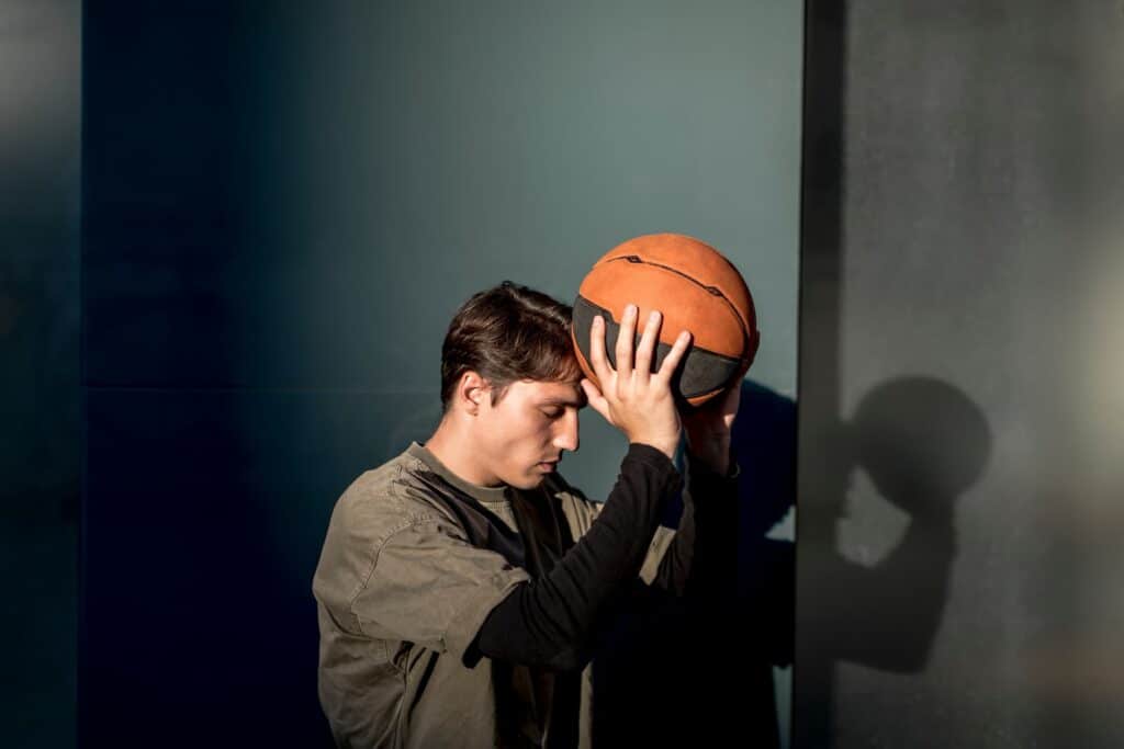 Un joven en postura pensativa, con los ojos cerrados y sosteniendo un balón de baloncesto contra la frente, apoyado en una pared azul grisácea con luz natural que crea sombras, reflejando concentración, pasión y reflexión deportiva.