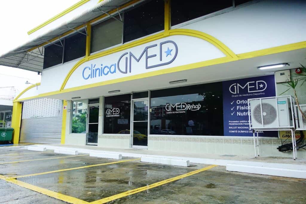 Fachada moderna de Clínica GMEDSport con ventanales, señalización de servicios y áreas de estacionamiento.