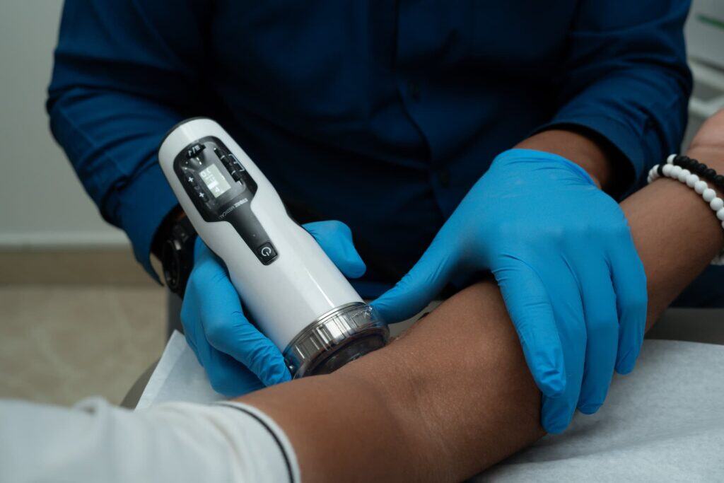 Una persona con guantes azules utiliza un dispositivo médico manual en el antebrazo de otra persona, posiblemente para examinar o tratar lesiones, en un entorno clínico.